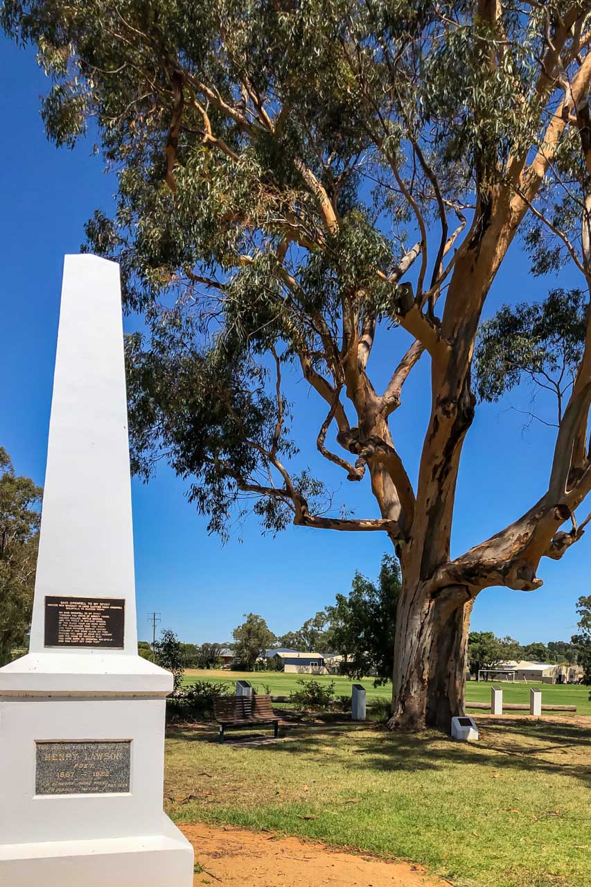 A large white obelisk. A tall gum tree, a wooden seat and plaques.