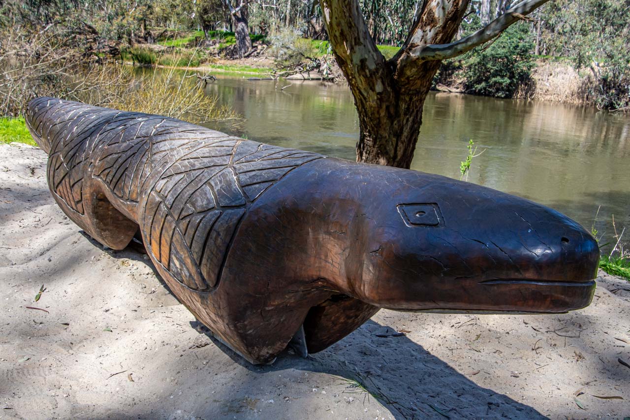 A photo of a large, carved wooden goanna, with a river behind the sculpture