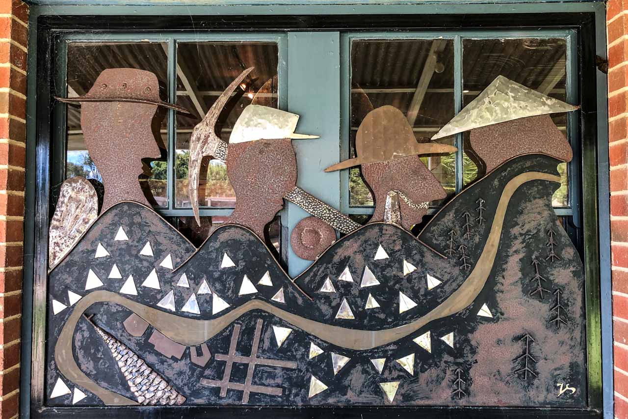 A flat metal sculpture in a window of four miners. The base of the sculpture depicts mountains and a river.