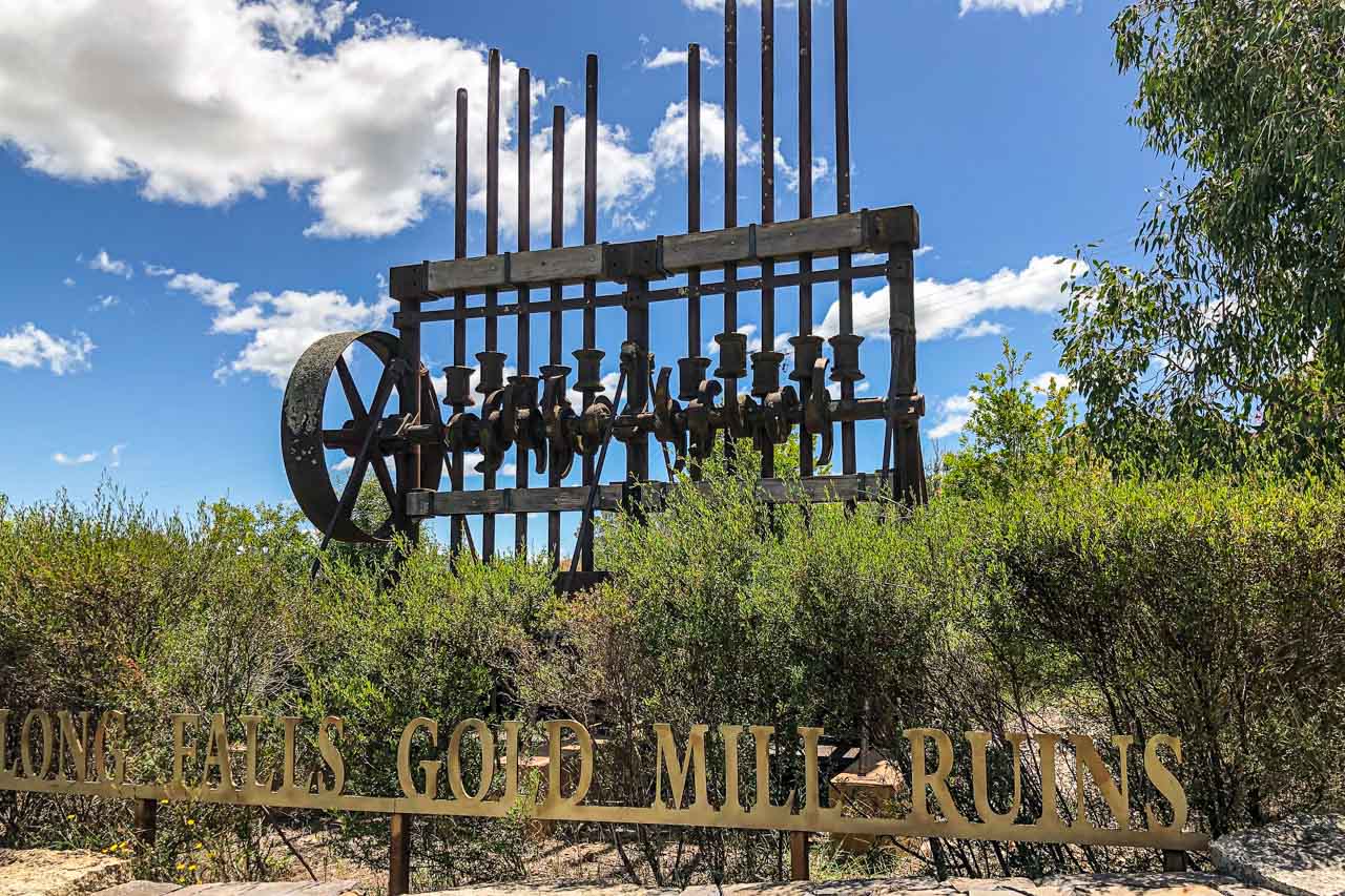 A large piece of gold mining machinery on the side of the road. The sign in front of the machinery reads, Adelong Falls Gold Mill Ruins