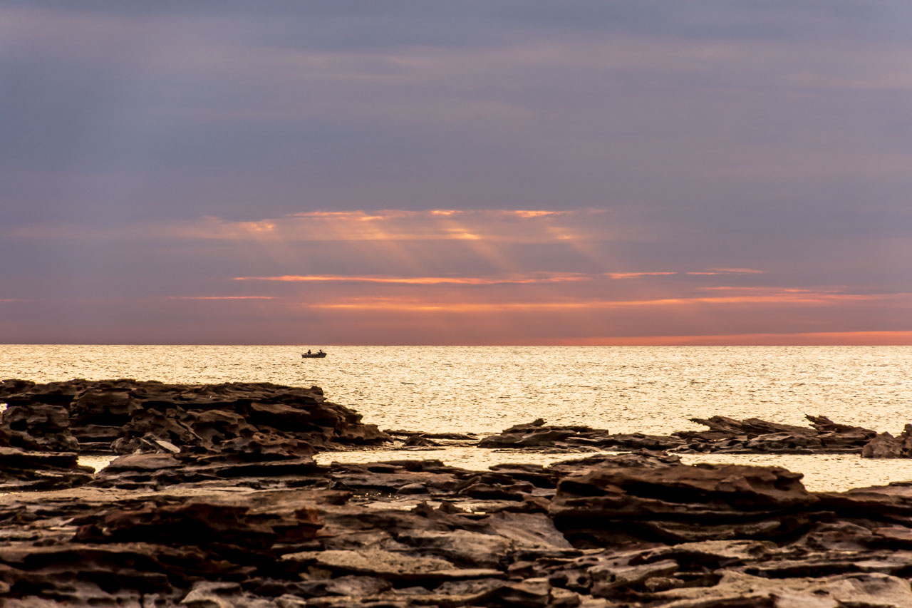 The sun sets over the ocean with rocks in the foreground. Two fishermen in a small boat are out to sea.