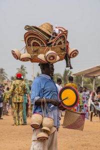 A man carrying straw baskets and hat on his head. He also has straw baskets hanging from his arm and carrying a straw fan.