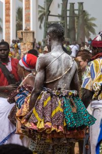A male Voodoo devotee dressed in a multi-coloured skirt walking through a crowd of people.
