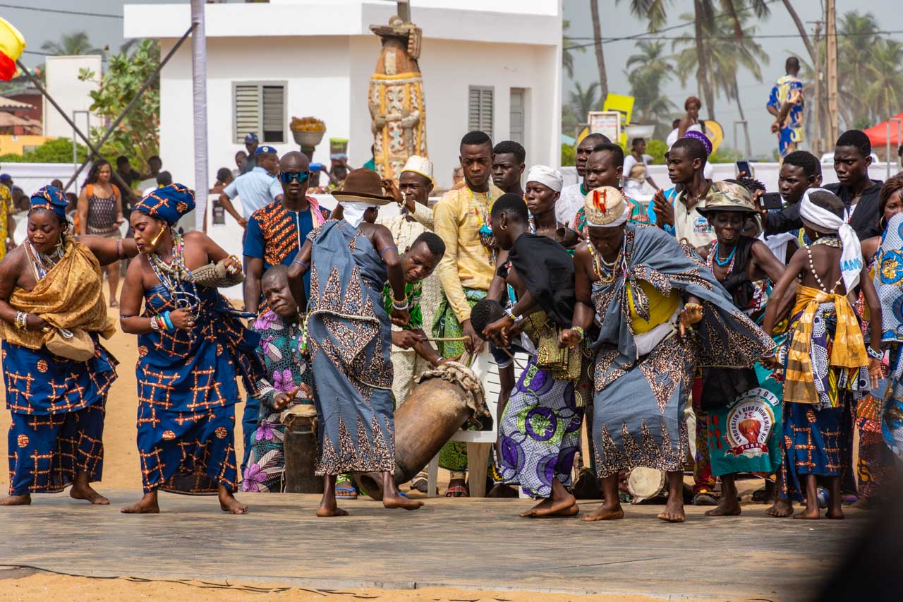 Men playing African drums and women dancing dressed in multi-coloured clothing. A crowd watches the drummers and dances.