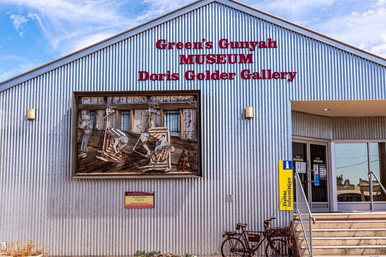 Corrugated iron building facade with a sculpture hanging on the front and a rusty bicycle near the entrance steps. The building is a museum.