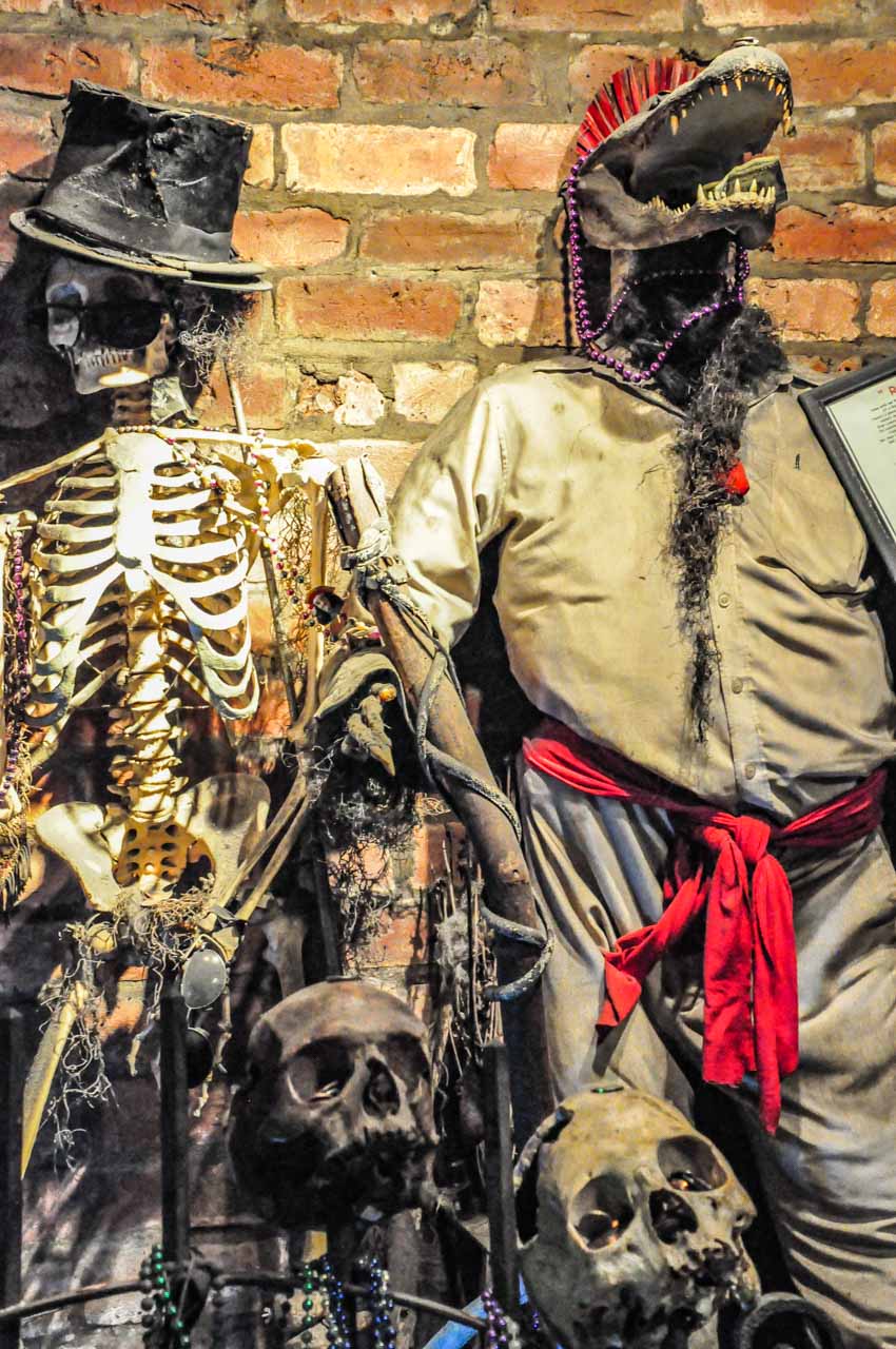 A picture of two human skulls, a human skeleton with a top hat, and stuffed clothes with an alligator skulls.