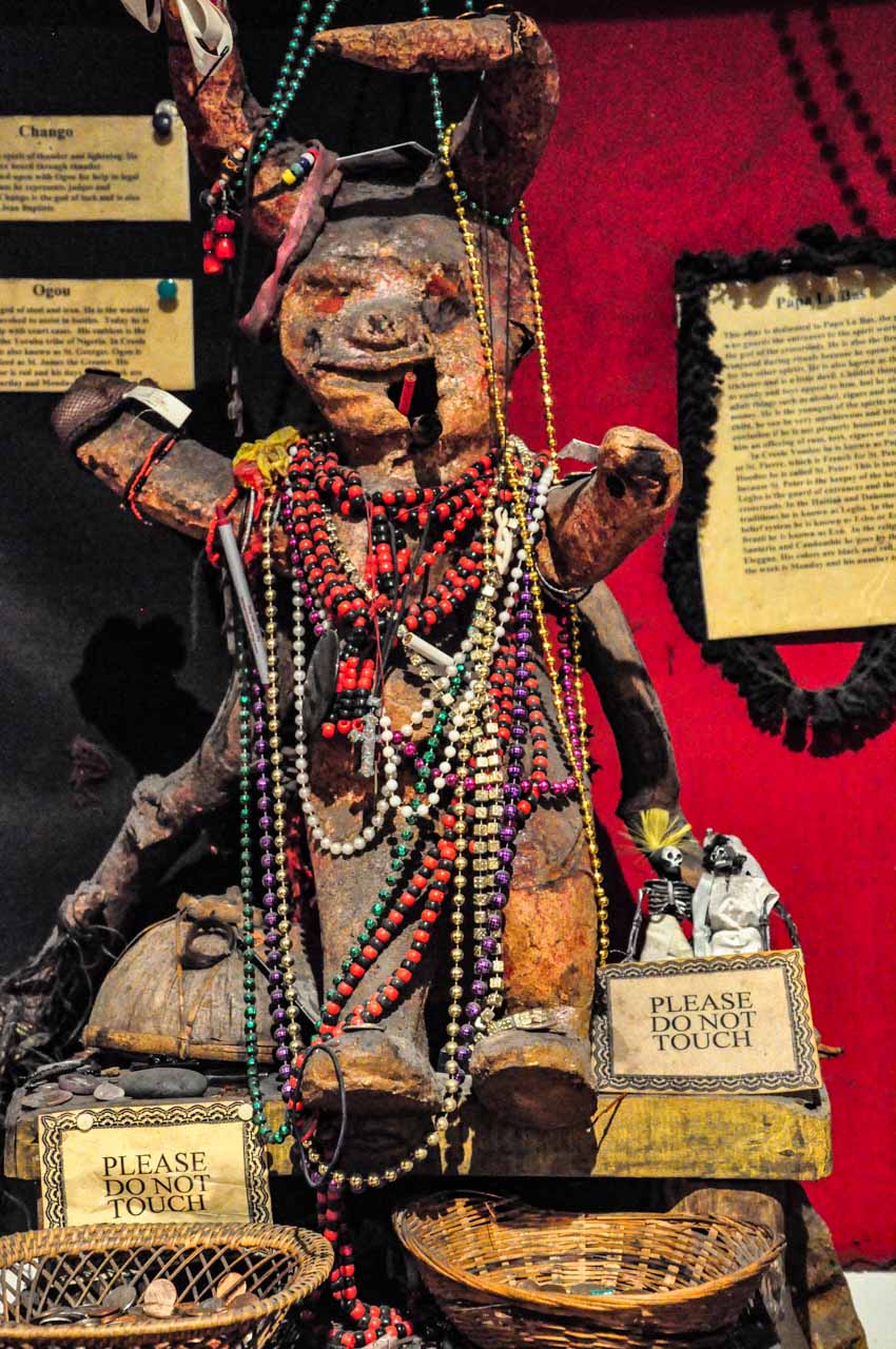 A clay 'human' figurine with horns on its head, and multi-coloured bead necklaces around it neck.