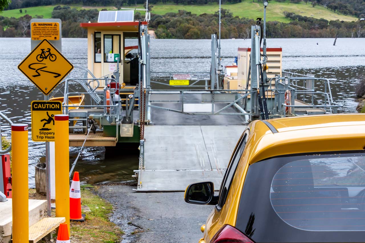 A yellow car waits to drive onto a three-car cable ferry to cross a river.
