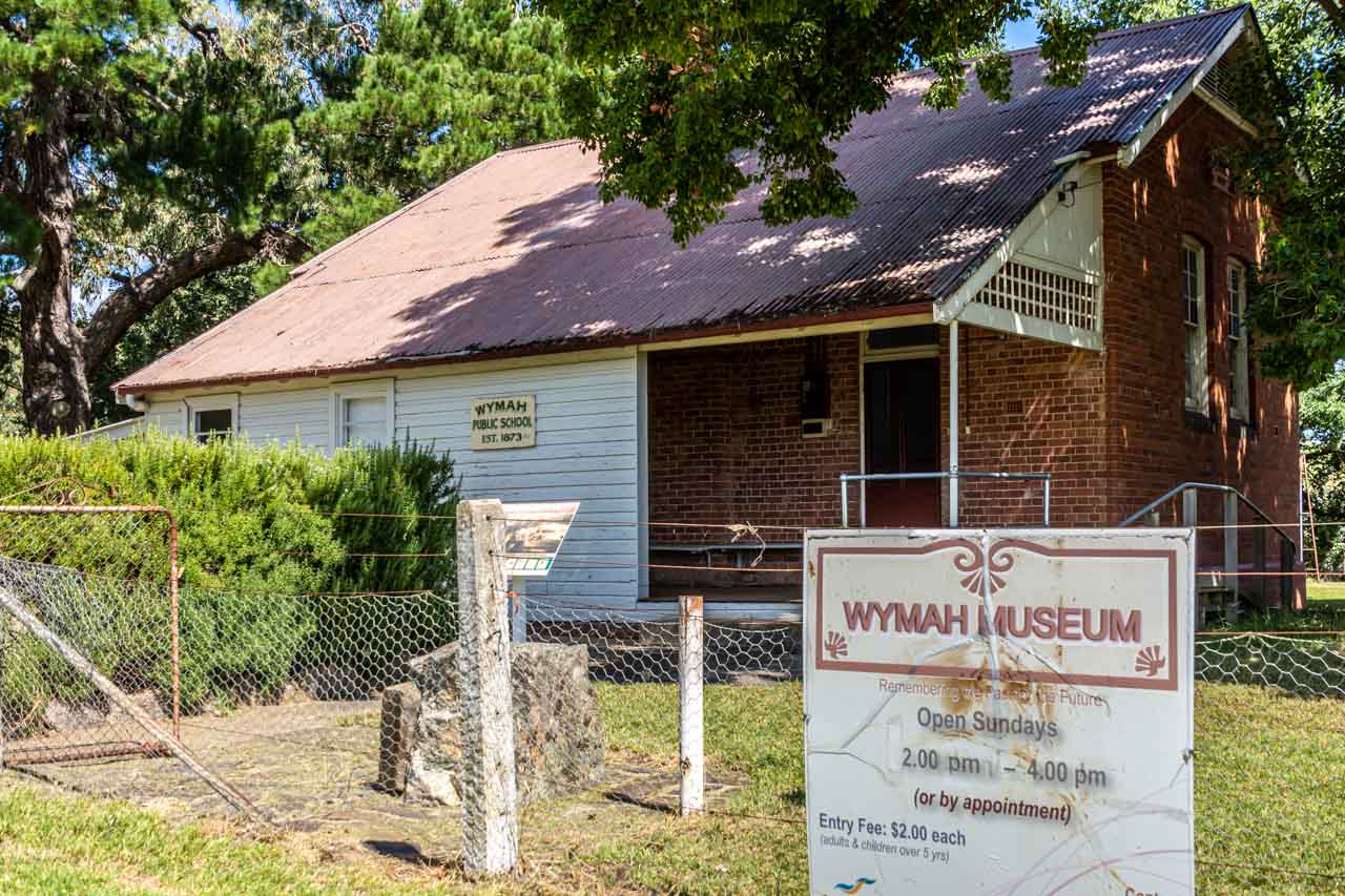 A brick and timber building with a red iron roof. The plaque on the building says, Wymah Public School, Est. 1873. The sign on the fence in from to the building says, Wymah Museum.