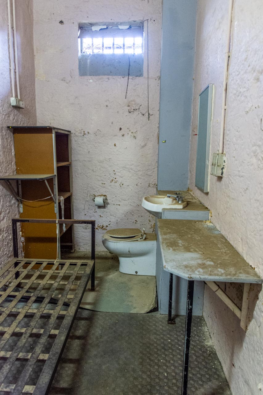A prion cell in an old gaol with toilet, hand basin, bench, cupboard, and iron single bed.