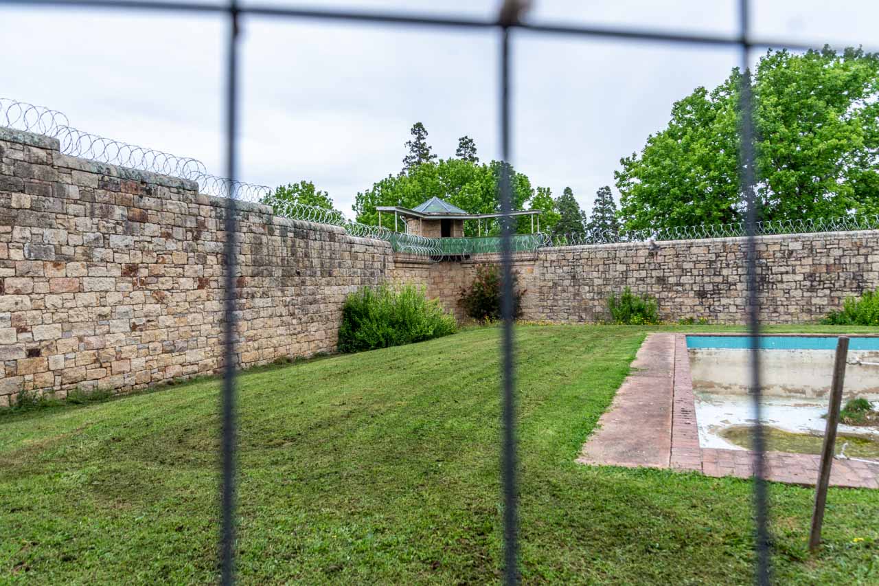 Image of a lawned area with empty swimming pool, surrounded by stone walls with razor wire and overlooked by a watch tower.