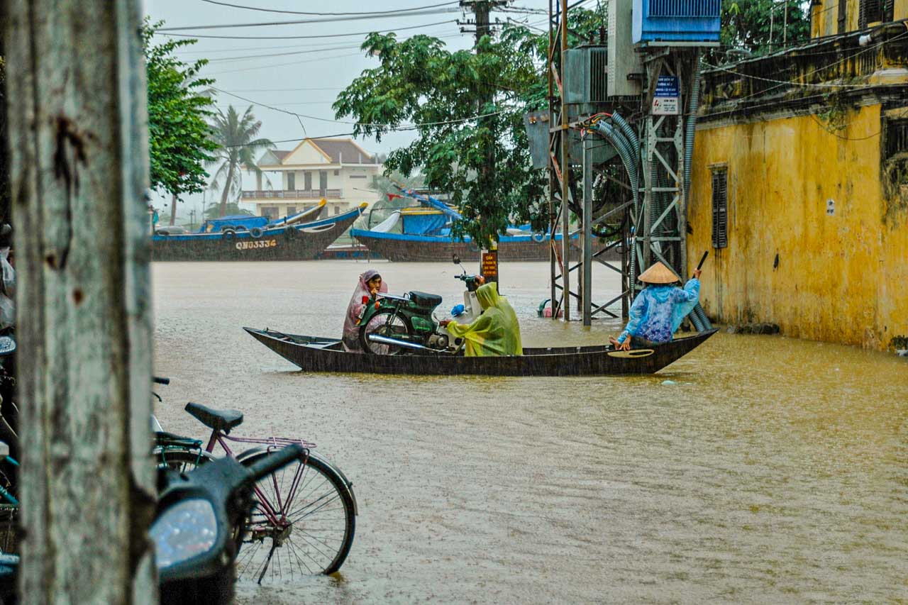 A photo of rain falling on a river that has flooded the street. People are transporting a motor bike in a boat along the flooded street.
