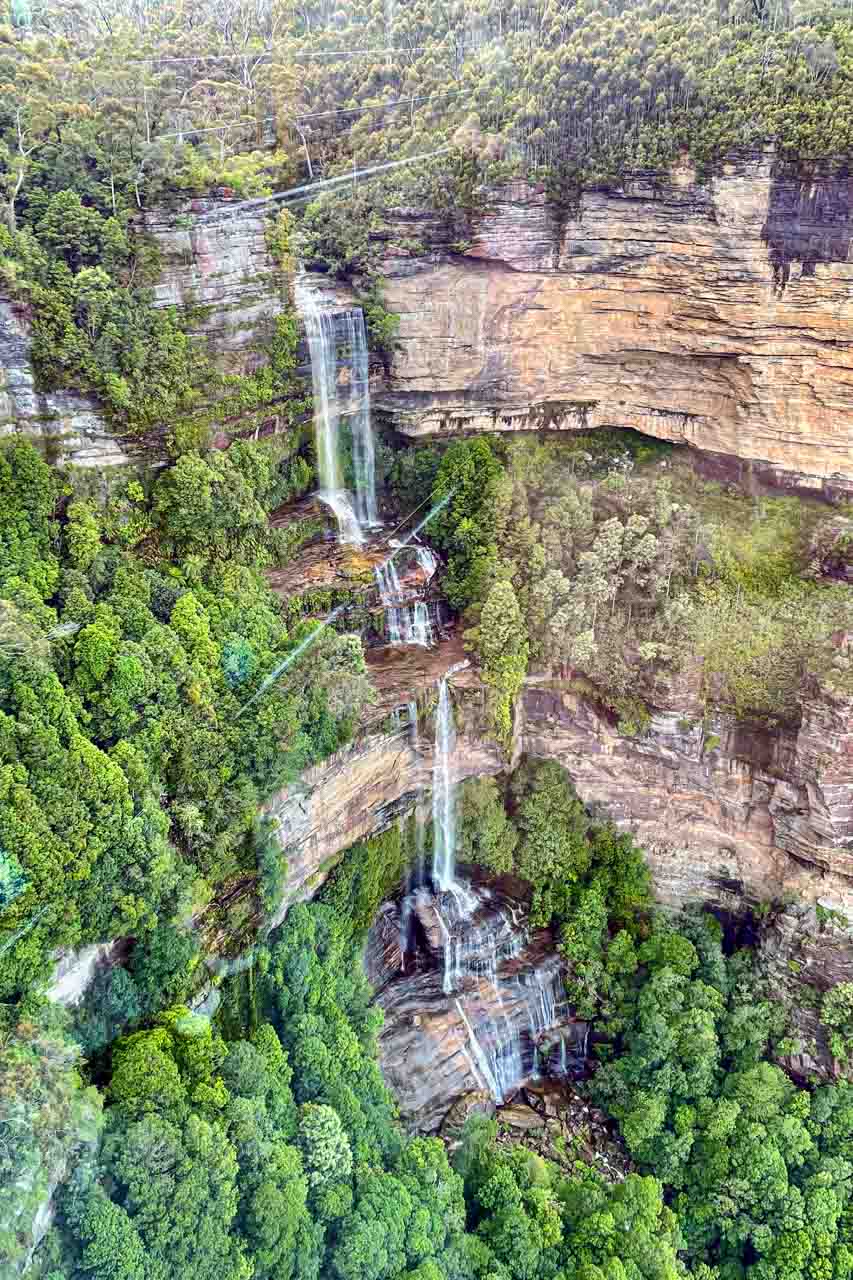 A photo of a multi-tiered waterfall plunging down a cliff face and surrounded by bushland.