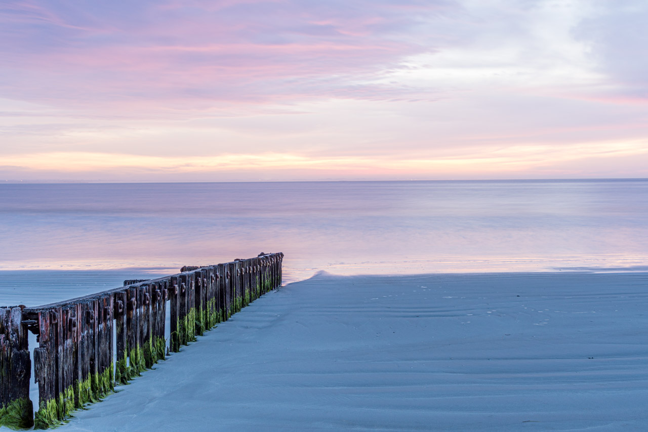 A photo taken at sunrise of timber fencing on the beach and leading into the ocean.