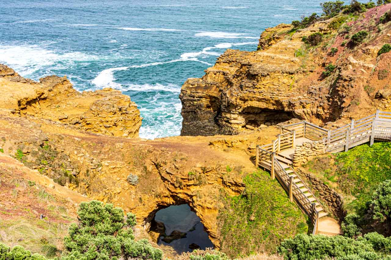 A picture of a steps leading down to a rocky coastline with the ocean in the background. One of the rock formations has an arch with a view to the ocean.