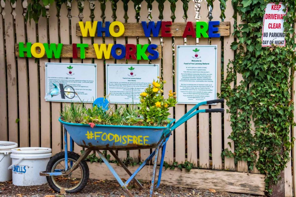 A photo of a blue wheelbarrow with plants in it and FoodIsFree written on the side. There is a wooden picket fence behind the wheelbarrow. On the fence is written, Who We Are and How It Works.