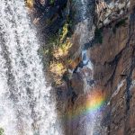 Photo of a waterfall with a rainbow reflected on the rock cliff face.