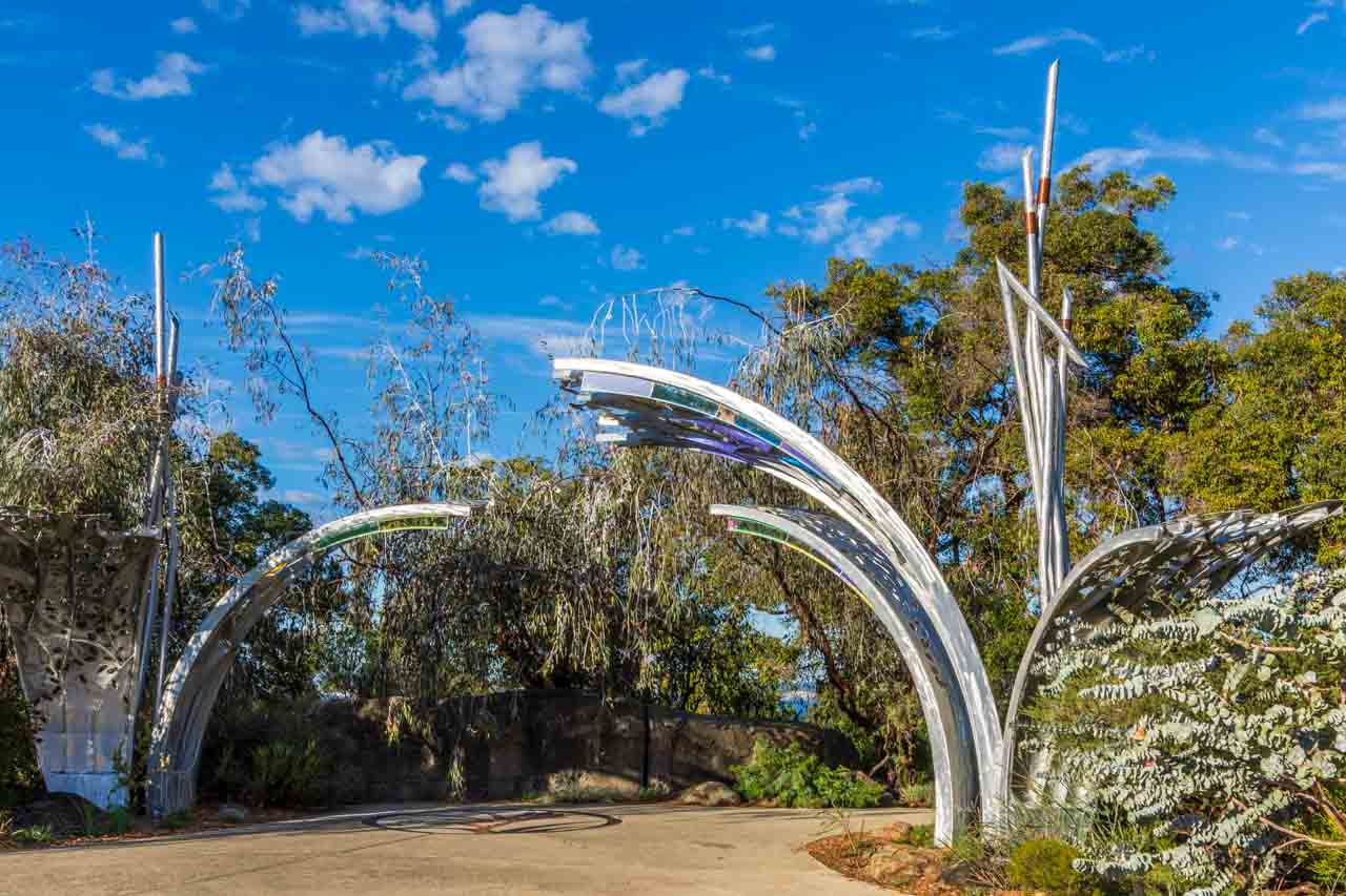 Curved silver metal sculpture overhanging a path and surrounded on two sides by shrubs