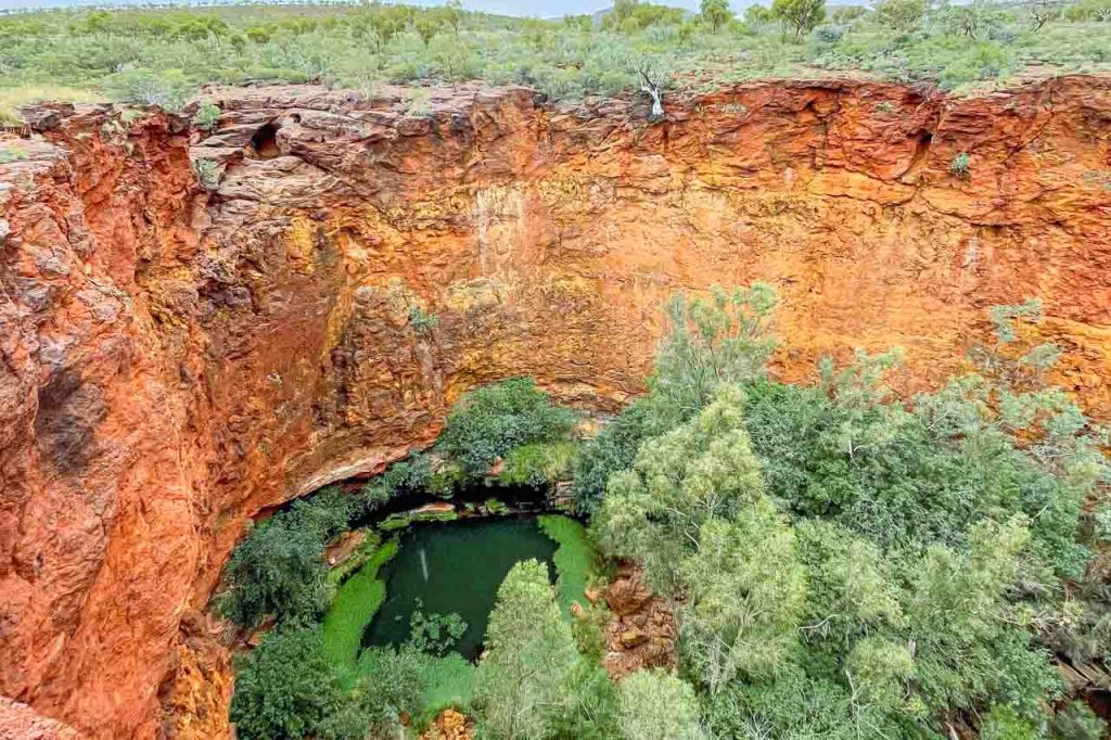 A pool of water at the bottom of a deep hole surrounded by red cliffs and trees