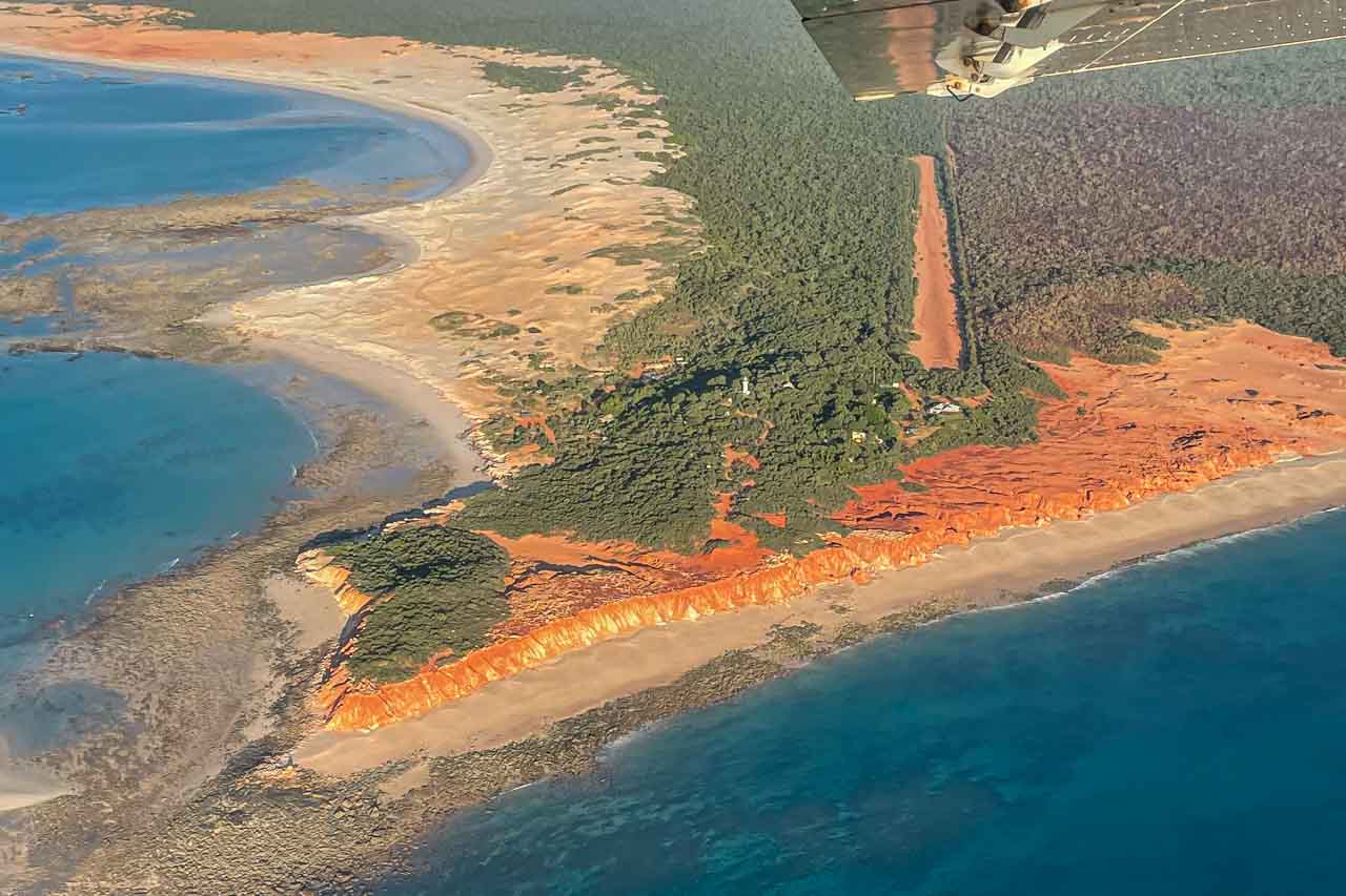 The wing of a plane is seen flying over a landscape of red cliffs, white sand, blue ocean and green forest
