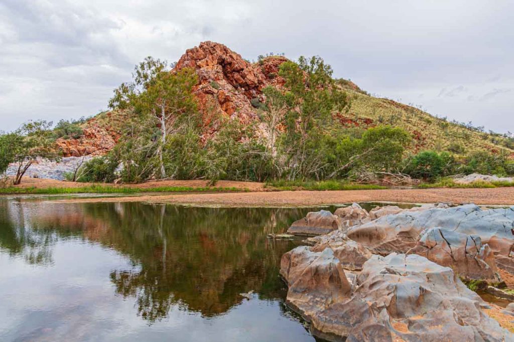 A red-coloured hill is reflected in a river with rocks in the foreground