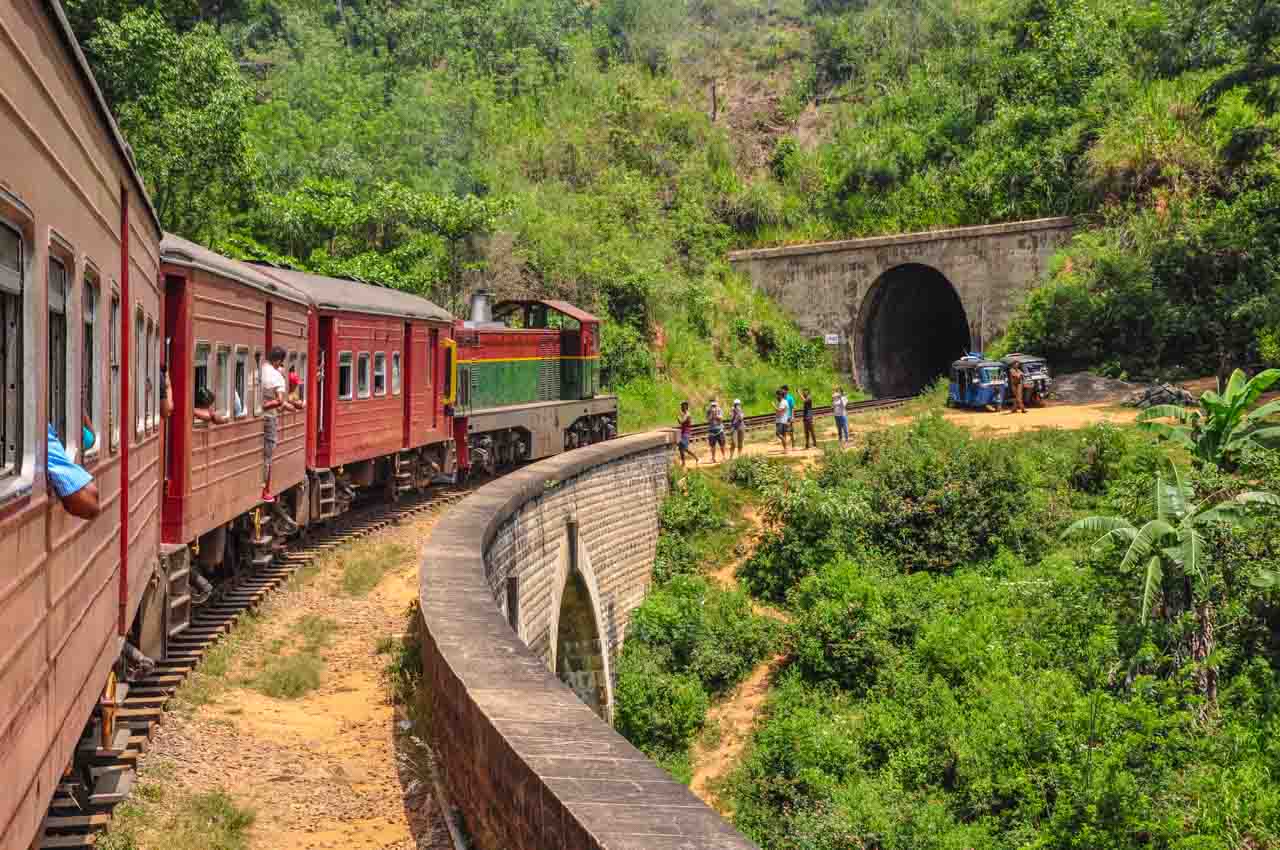 A train on a bridge approaches a tunnel and people standing near the bridge wave at the train.