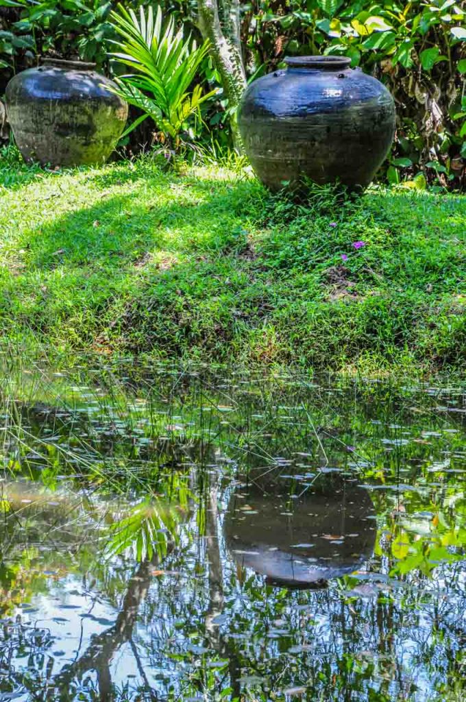 A large black jar sitting on grass is reflected in the waters of a pond