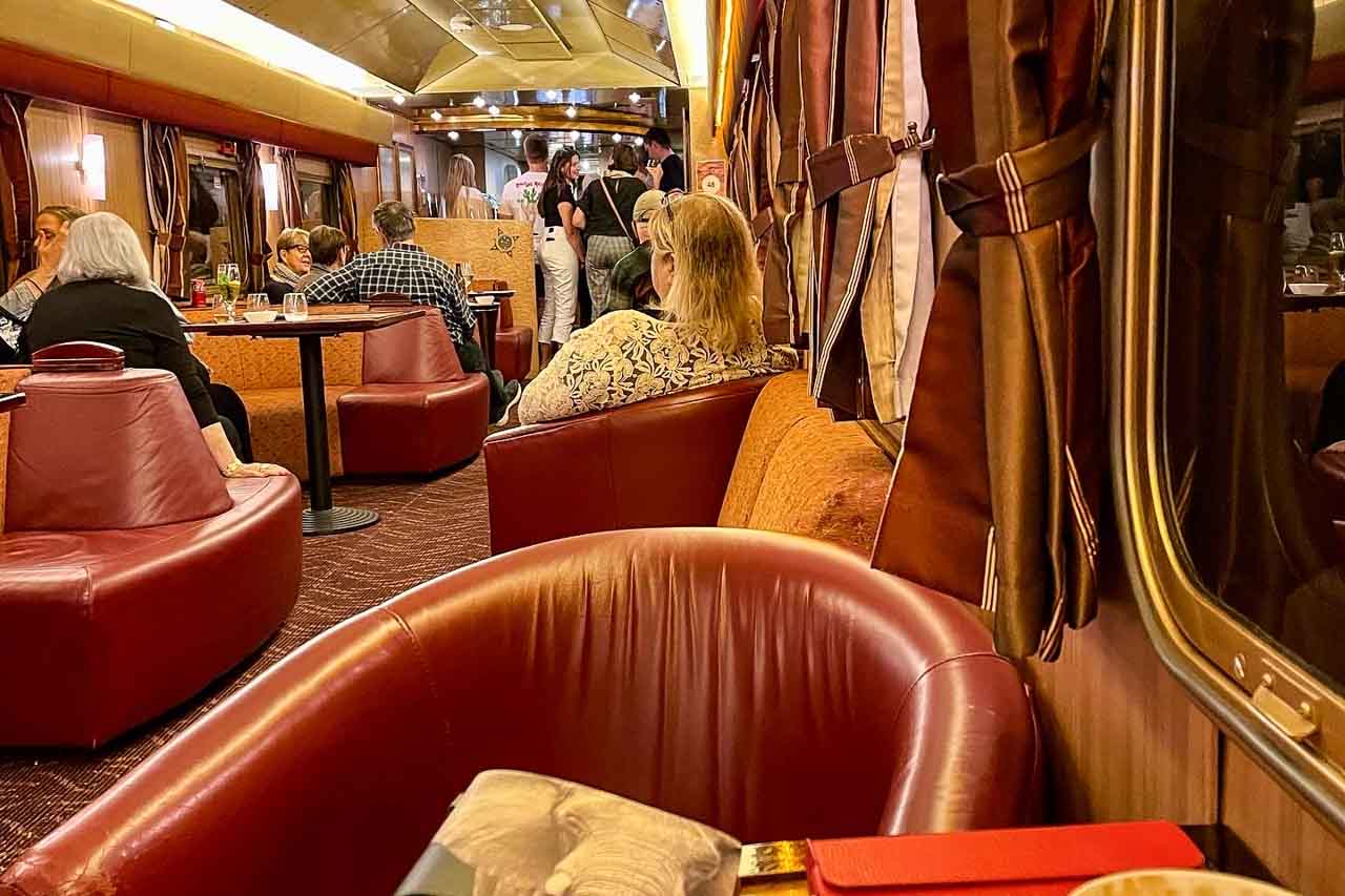 A lounge train carriage with tables, leather and material seats and couches. People are sitting on the seats, and drinks are on the tables.