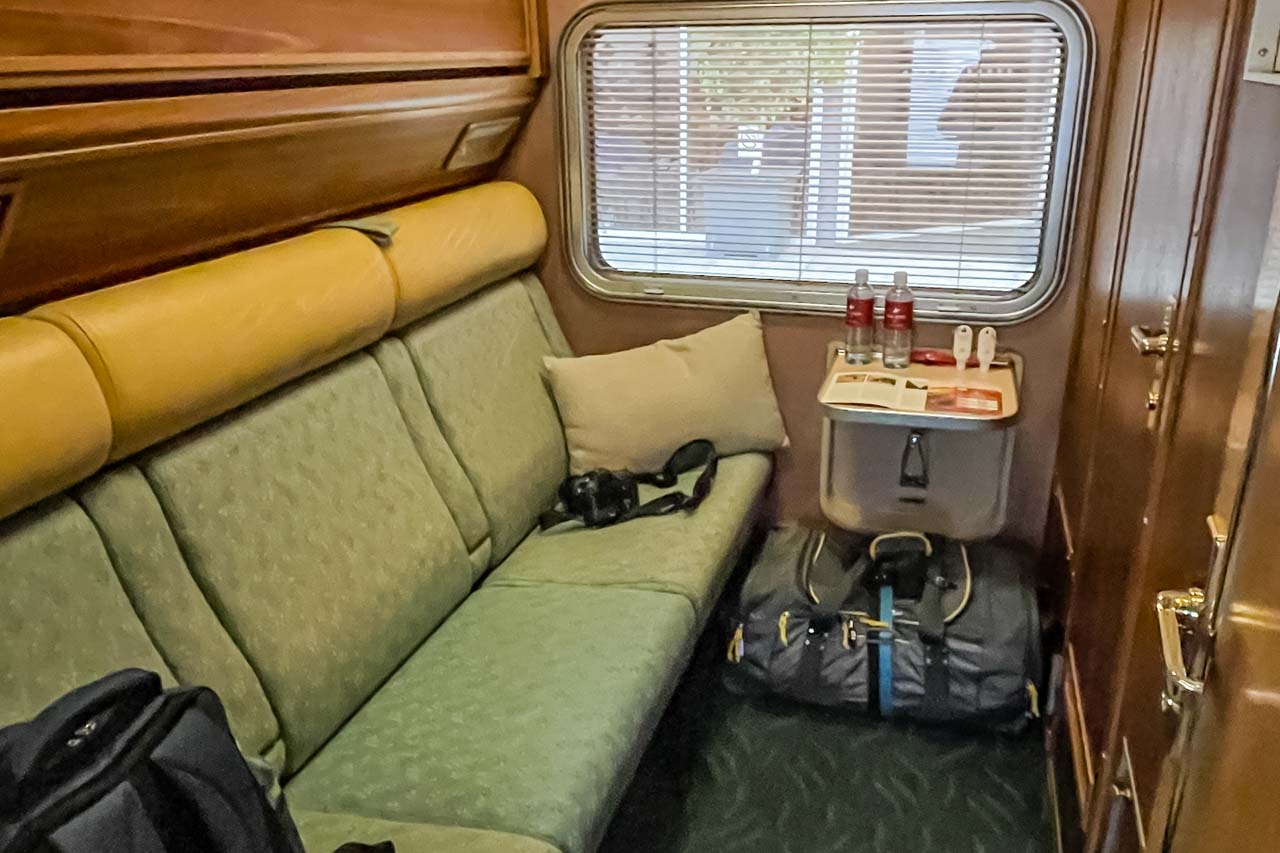 A train cabin with a three-seater couch. A bag and camera are on the couch. The door handle to the ensuite can be seen and there is a bag on the floor. Water bottles are on a small table attached to the cabin wall.