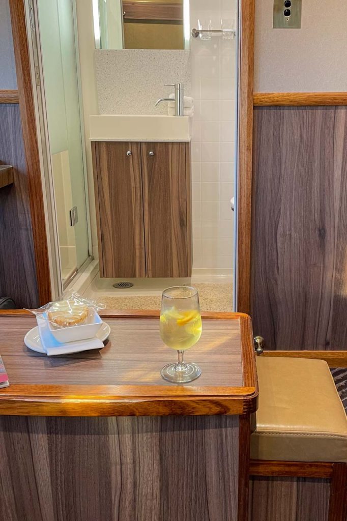 The photo is a view of a train cabin ensuite. In the foreground is a table with a drink and biscuits on it.