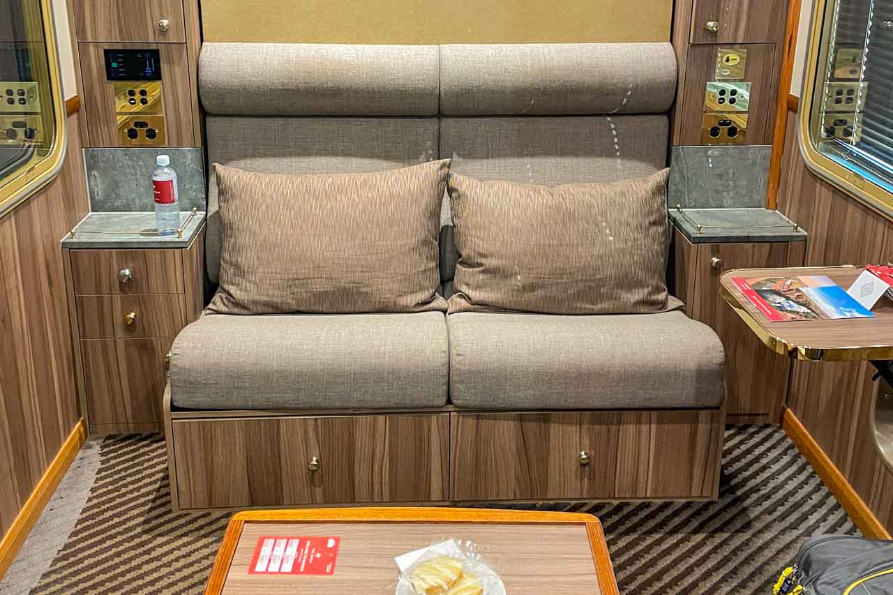 A train cabin with a two-seater couch with cushions. There are cupboards each side of the couch and two tables - one is in the middle of the cabin and one is attached to the cabin wall.
