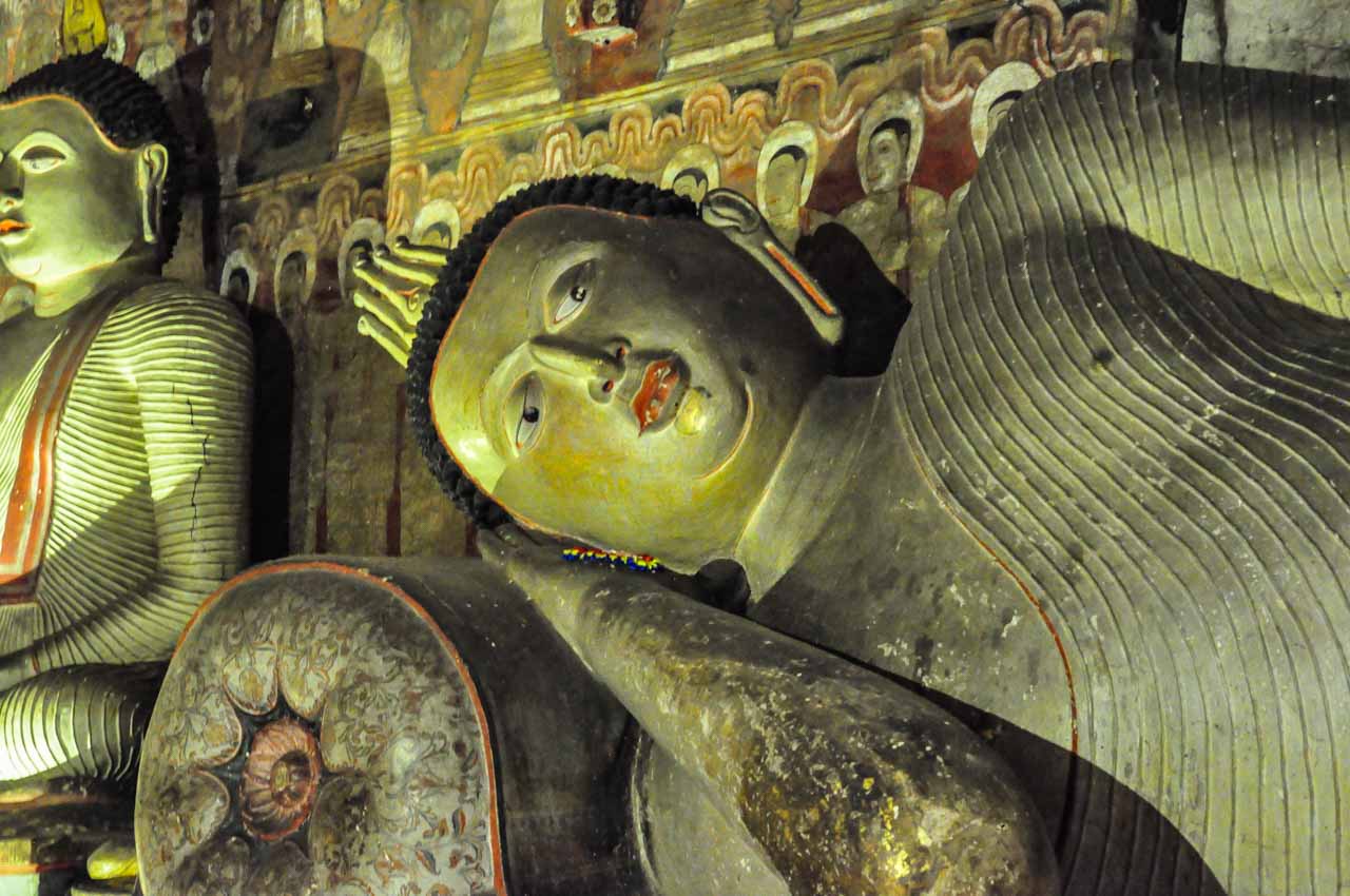 A partial view of a reclining Buddha and sitting Buddha with frescos on the wall behind them.