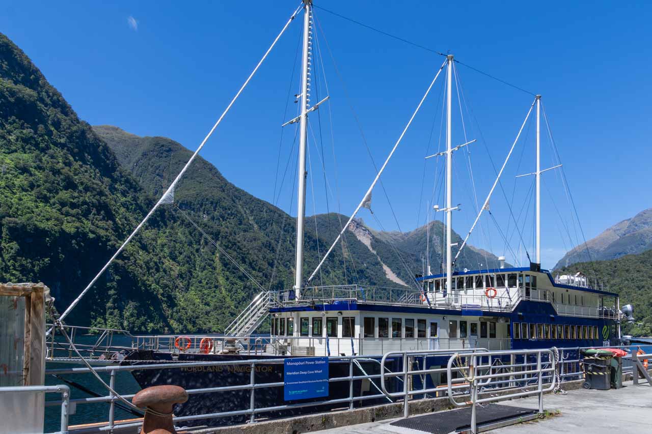 A three-mast passenger ship docked at a wharf, with mountains in the background.