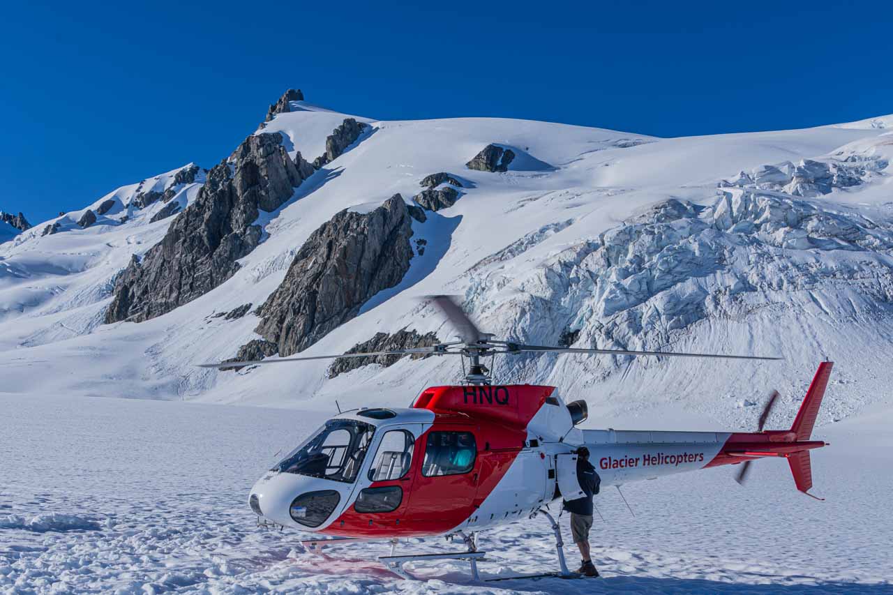A red and white helicopter sits on the snow with mountains behind it. The name, Glacier Helicopters is printer on the helicopter's tail. The pilot is looking into the helicopter through an open door in the tail section.