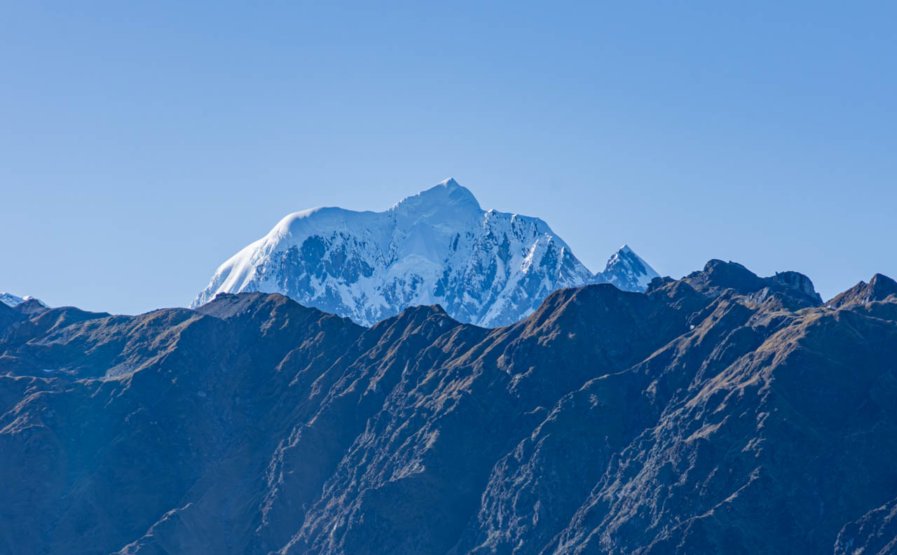 A snow capped mountain rises above a mountain range devoid of snow.