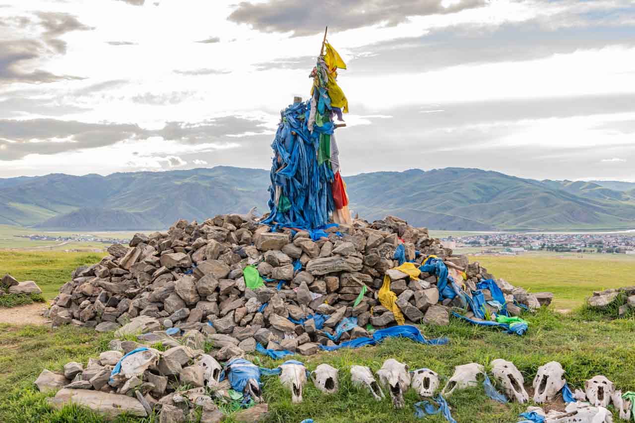 A pile of rocks and stones with a pole in the middle of the mound covered in blue scarfs. Animal skulls are placed in front of the mound.