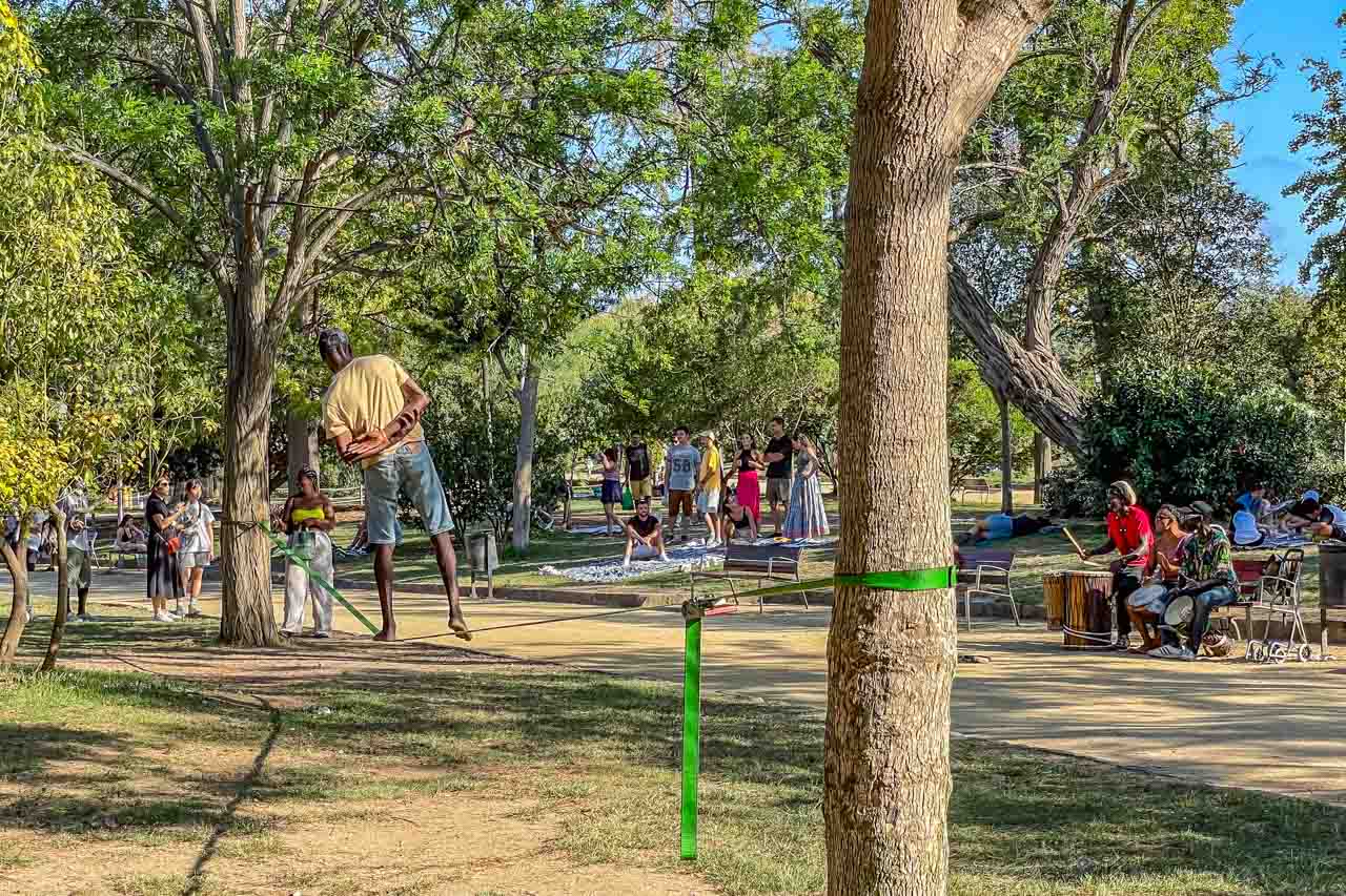 A man walks a tight rope slung between two trees while drumers play and people watch.