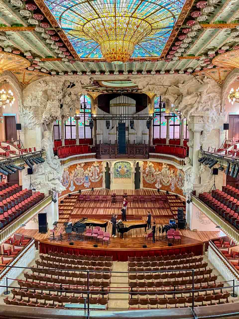 The interior of a music hall with an organ, muses on the back wall of the stage, a piona on the stage, a stained-glass ceiling, and seating on two tiers.