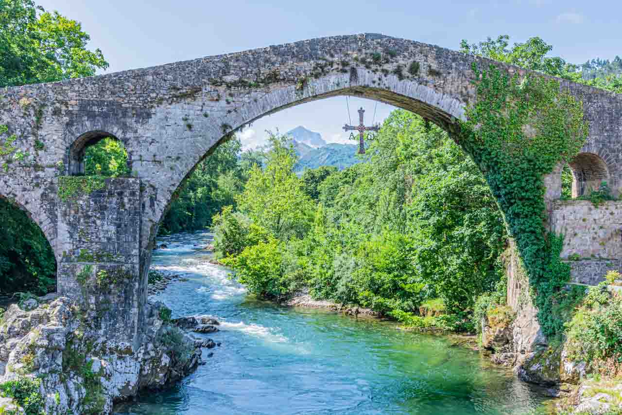 An arched stone bridge with a metal and jewelled cross hanging from the middle arch spans a river.