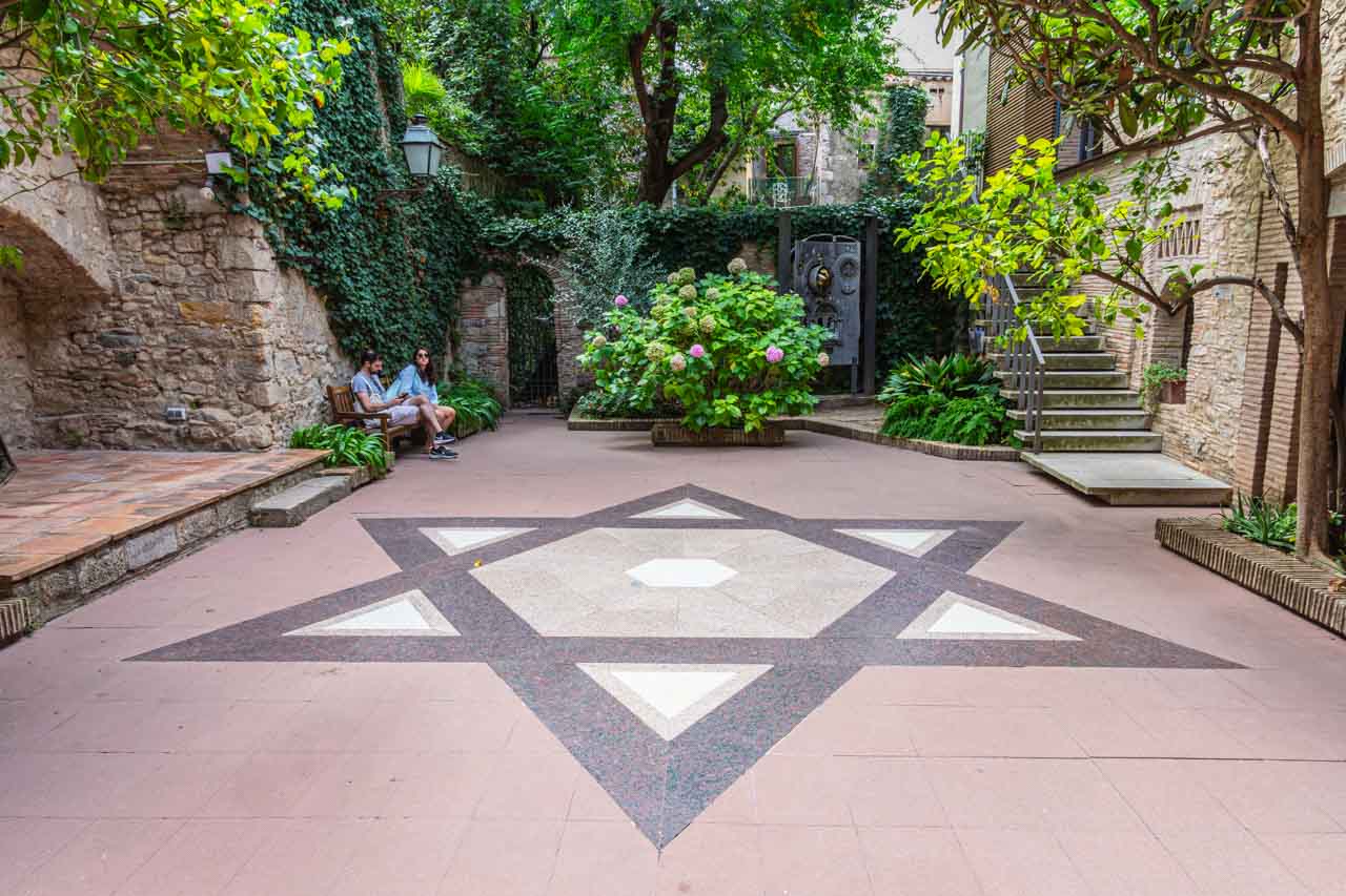 A Jewish Star of David i n tiles is incorporated in a tiles courtyard. Two people sit on a bench in the courtyard. There are flowers and shrubs at the end of the courtyard.