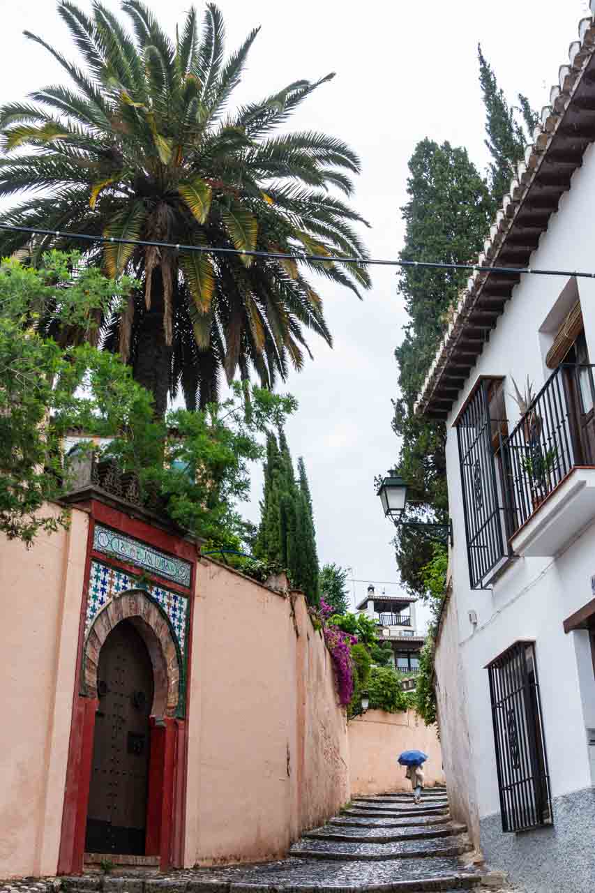 A stepped street with Moorish architecture (building on the left) and an Andalusian styled building on the right. A lady with a blue umbrella is walking up the stone steps.