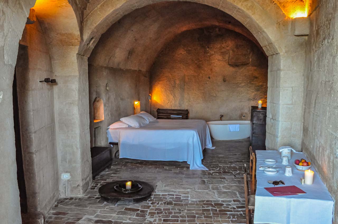 A hotel room in a cave, with a double bed, bath, and table and chairs. the cave room is lit by candles.