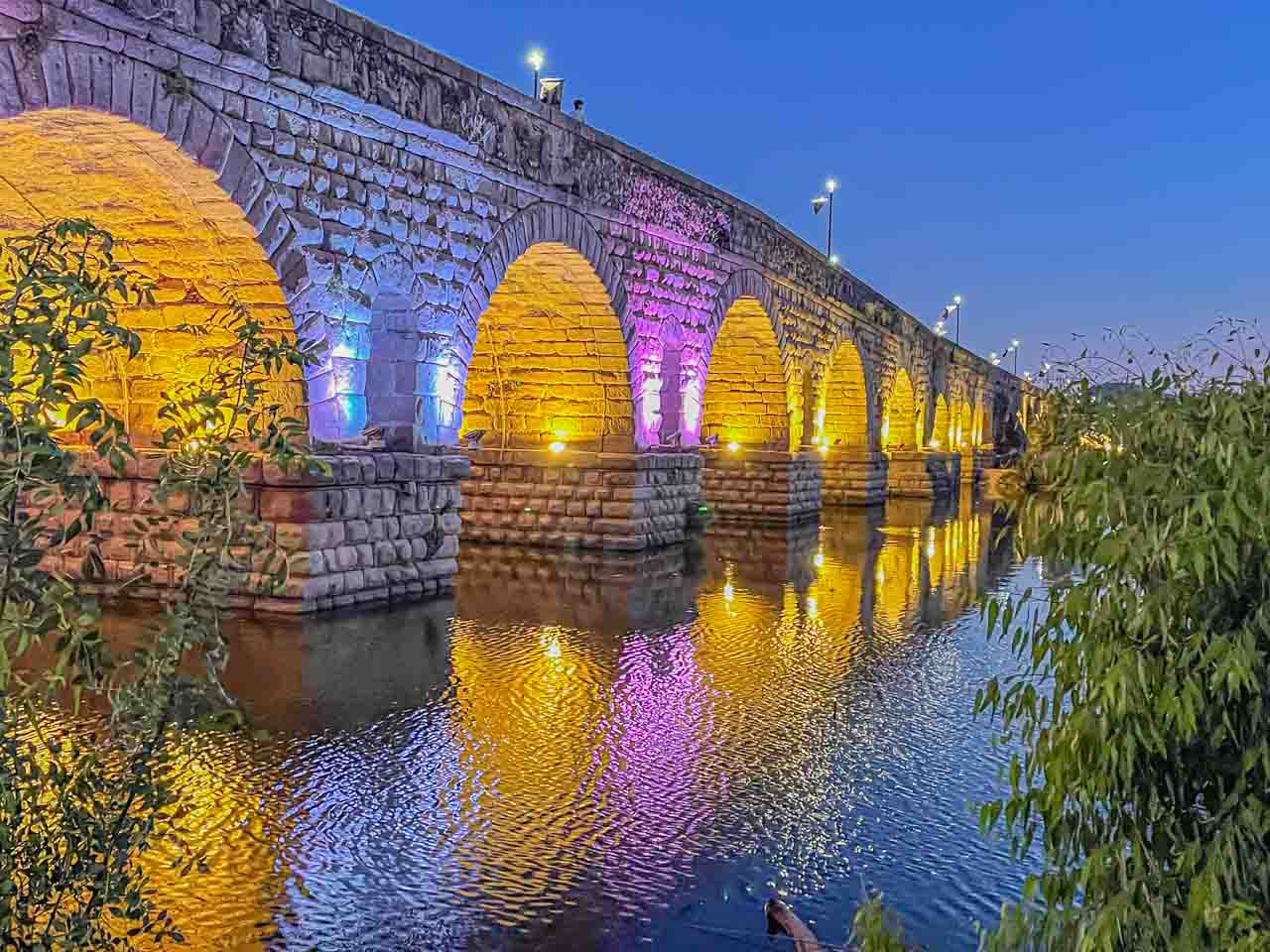 A stone bridge built by the ancient Romans is lit with blue, pink, and yellow lights which are reflected in the river.