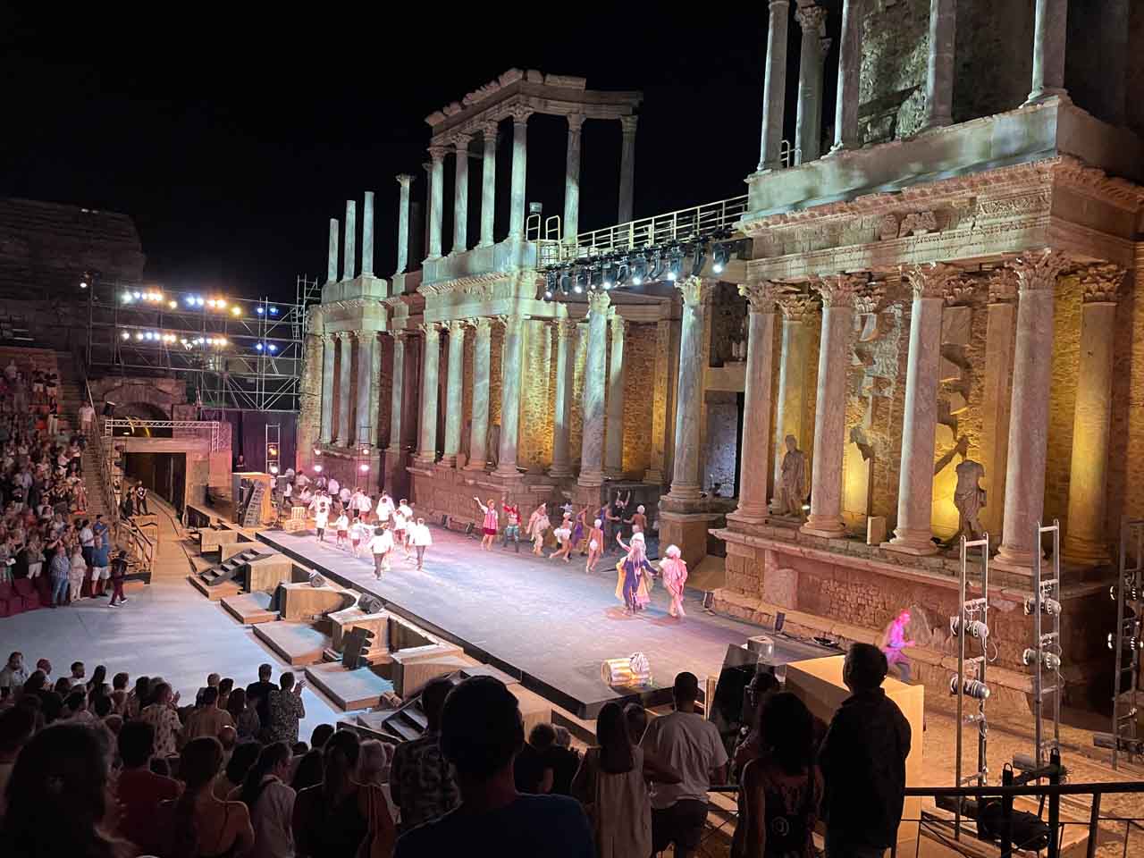 People standing and clapping the actors on the stage of a Roman theatre. The photo is taken at night with the stage lit up.