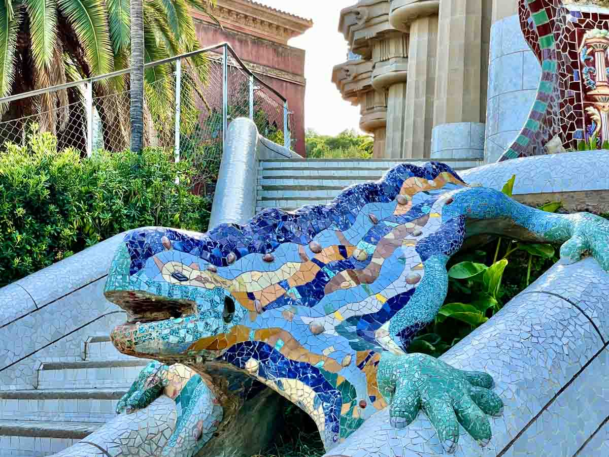 A blue, green, and brown mosaic-covered lizard sculpture positioned between two flights of blue mosaic-covered stairs.