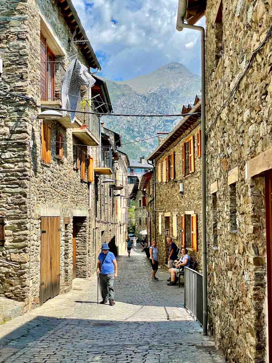 A man with a walking stick walks up a cobble stoned street with stone buildings on either side of the street.There are three other walkers with backpacks in the street.