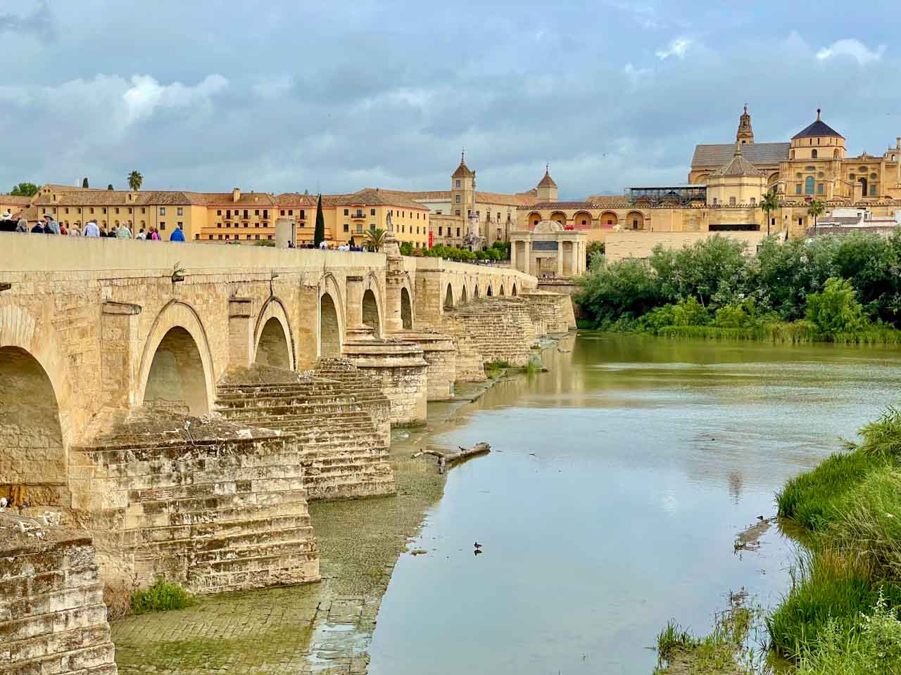 A multi-arched stone bridge with buttresses spans a river and leads to a medieval city.