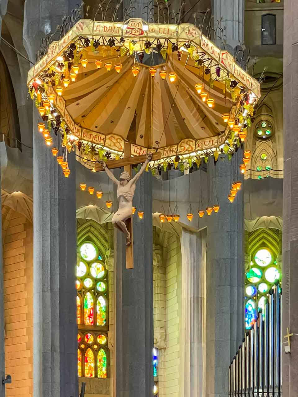 A statue of Jesus on a cross underneath an umbrella hangs fro the ceiling of a church. Grey marble columns and stained glass windows feature below the sculture of Jesus.