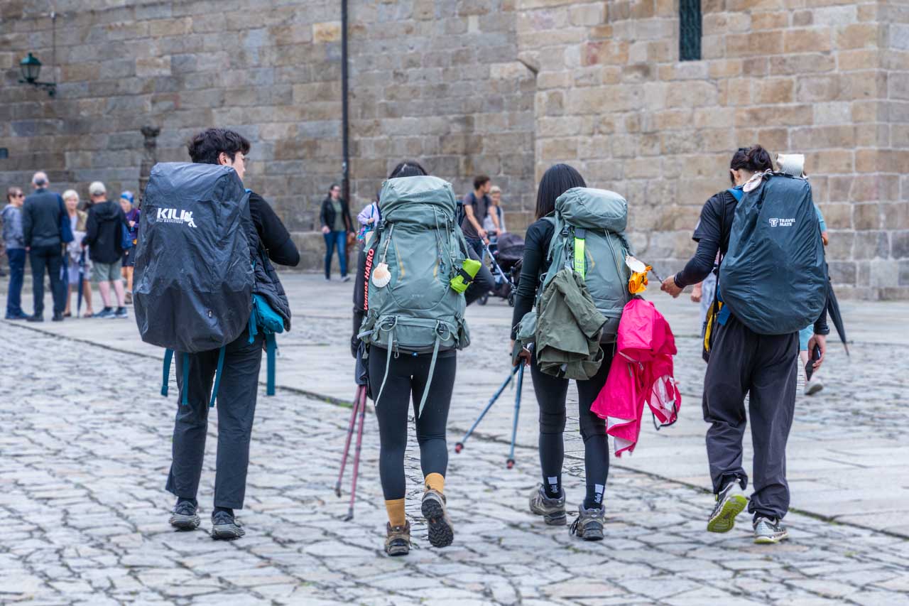 Four young people with backpacks on their backs and walking poles in their hands walk away from the camera on a cobblestoned street.