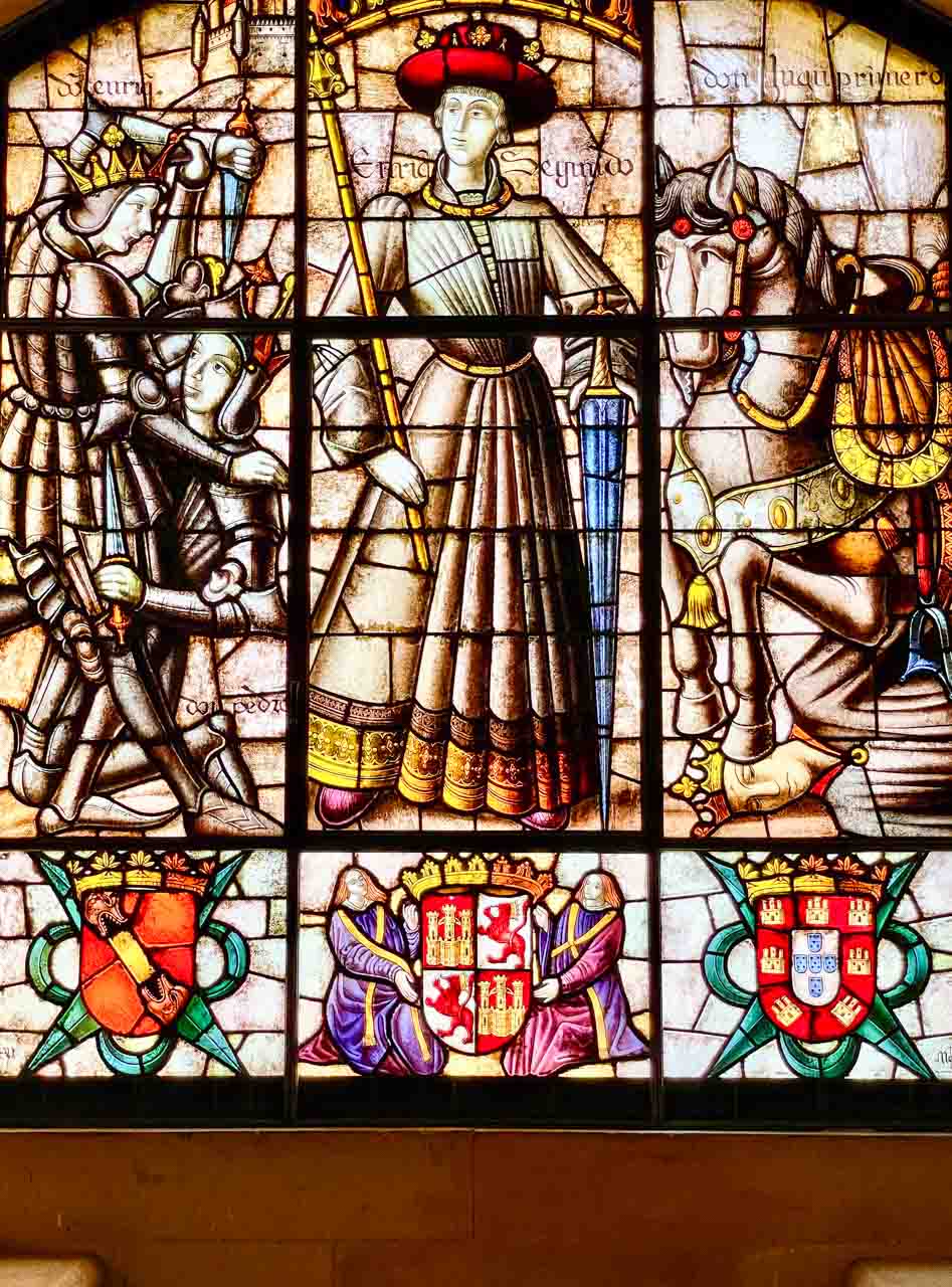 A stained glass wind depicting fighting medieval knights, a horse, a man in long robes, and three shields.